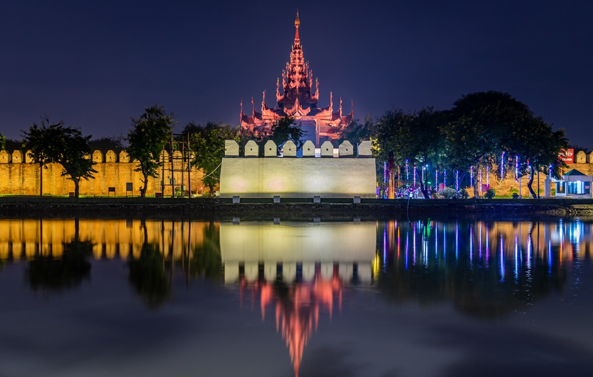 Mandalay Fortress at night — The walls and a watchtower of the Mandalay Palace fortress lit up at night, reflected in the still water of the moat.