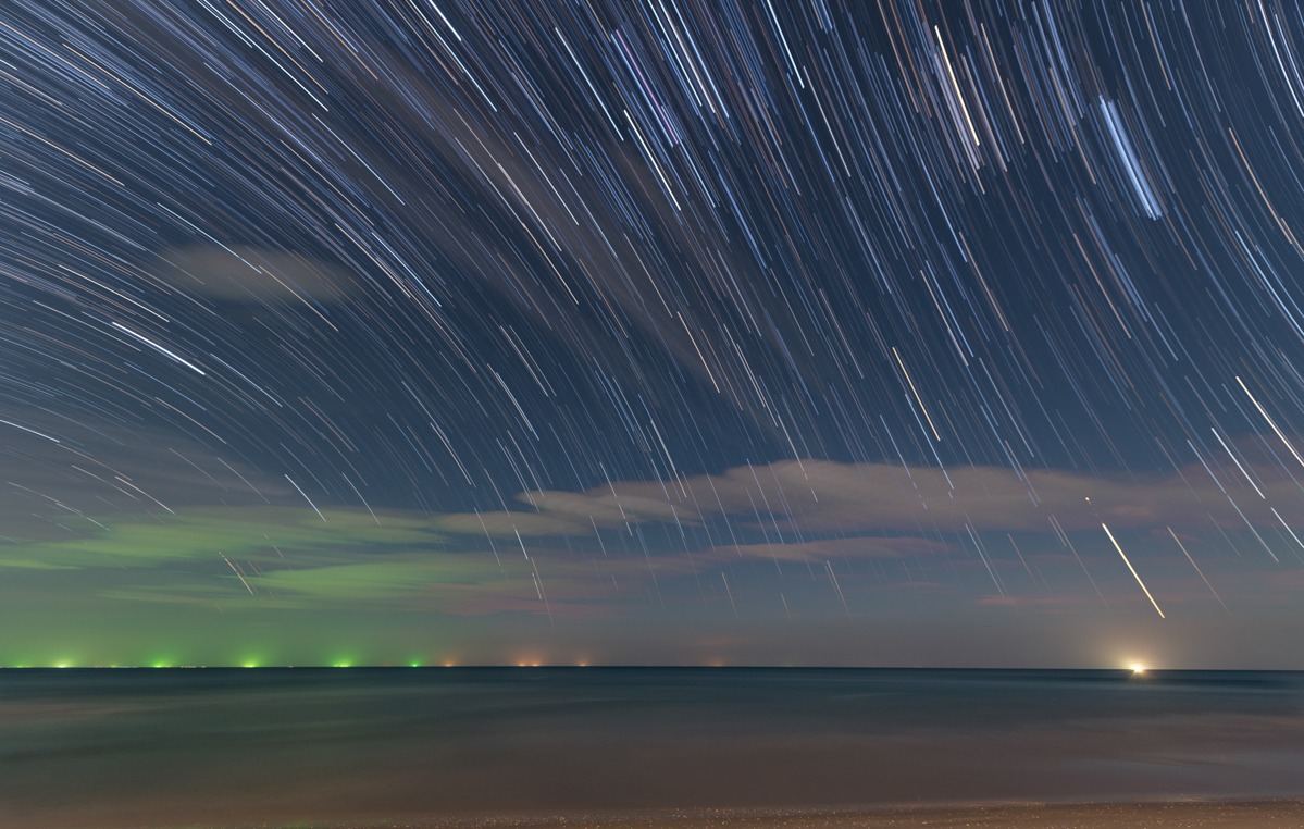 Star trails over the sea at night — Stars streaking across the night sky above a calm sea, with fishing-boat lights on the horizon.