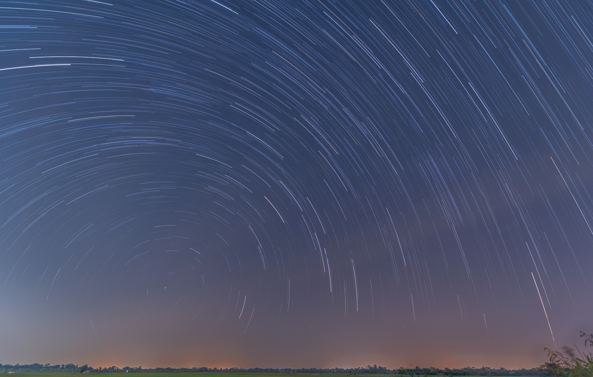 Circular star trails over open fields — Star trails circling the celestial pole above flat farmland, shot as a long sequence of exposures and stacked.